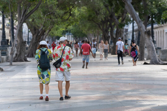 Older  couple of tourists in bright clothes and hats walking on Prado boulevard in the city center of Havana, Cuba. Selective focus, blurred background. Vacation, travel, tourism concept. 