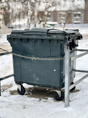 Urban Winter Scene With Trash. Snowy Neighborhood With Abandoned Garbage Container. Chained Rubbish Bin In Snowcovered Courtyard With Blurred Residential Buildings In Background