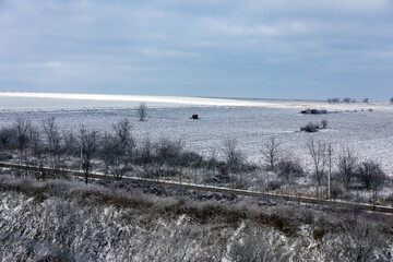 Wide view of a frosty rural field with sparse trees, a distant hut, and cloudy sky, capturing a tranquil winter scene ideal for seasonal and agricultural themes.