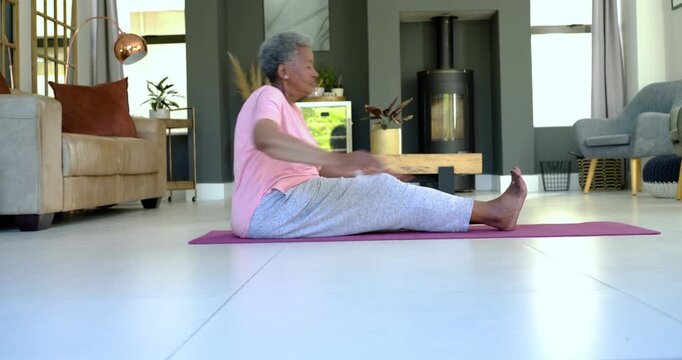 African American senior woman starting home routine, stretching legs and twisting torso on pink mat