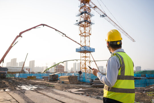 Construction worker wearing safety helmet and reflective vest checking smartphone on site with crane and concrete pump in background