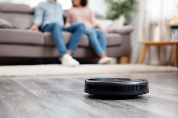 Robot vacuum cleaner cleaning a wooden floor of the house while a couple is resting at couch in home.