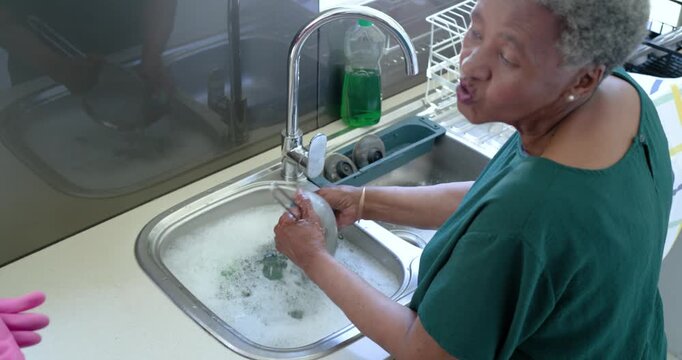 Senior African woman in green top scrubbing pan at soapy sink to clean, pink-gloved helper nearby