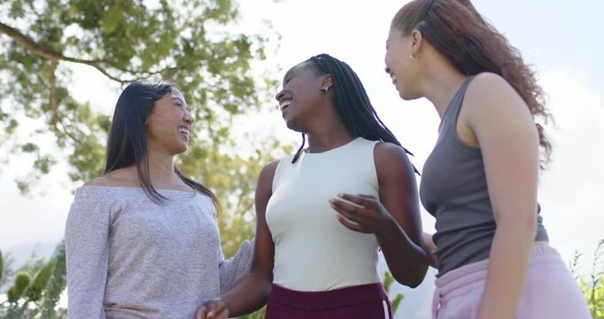 Diverse female friends smiling, making eye contact and leaning in, laughing and posing under tree