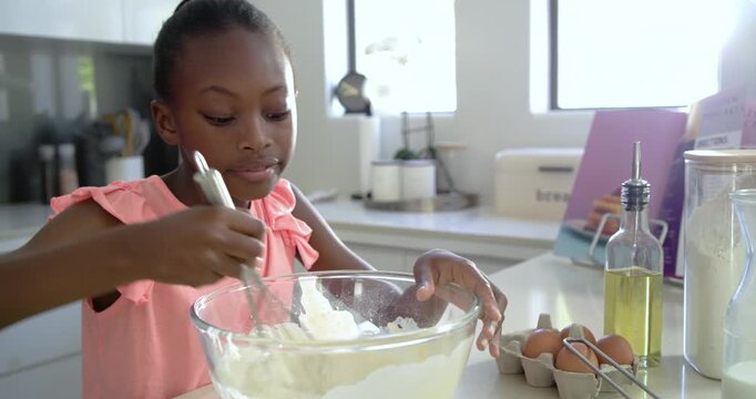 Cookbook guiding African American female child whisking batter with whisk at counter, checking
