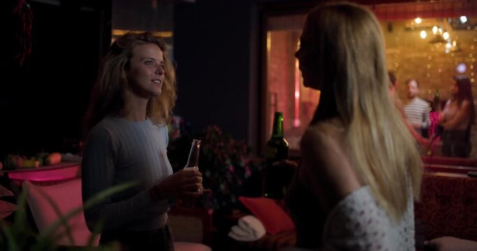 Friends talk and enjoy drinks inside a bar while people dance outside at night in a busy urban area