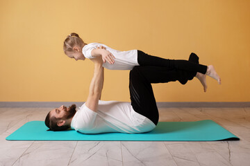 Happy father and young daughter practicing acro yoga on a mat indoors, playful bonding and healthy lifestyle concept, yellow background.