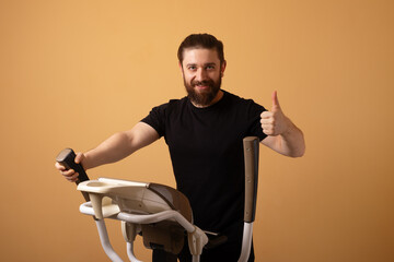 Smiling man in black shirt using elliptical machine and showing thumbs up, isolated on beige background, promoting healthy lifestyle and fitness.