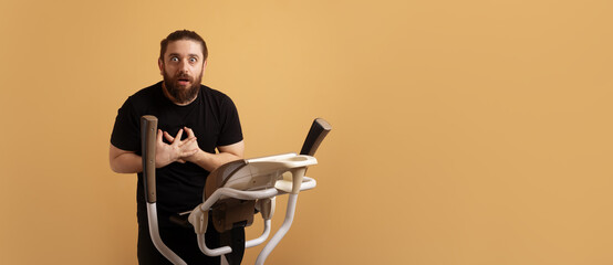 Shocked man in black shirt holding chest while standing on elliptical trainer against plain beige background, wide space for text.