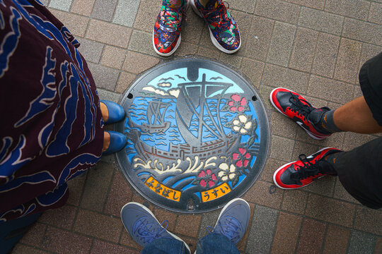 Tokyo, Japan - Oct 16 2024, close-up view of a manhole decorated with image of two ships on sea and camellia flowers, surrounded by feet of a family of four, Funabashi subway station, Tokyo, Japan