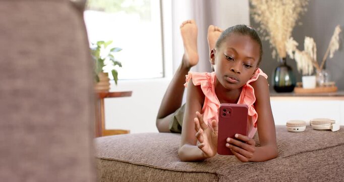 African teenage girl lying on ottoman near window, holding red phone, wearing pink top and reading