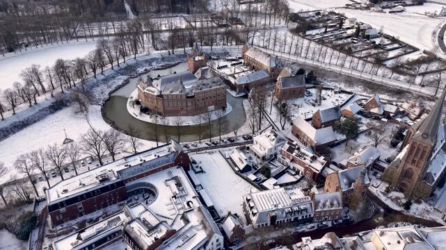 Aerial view of a snow covered historic town with a large brick castle and moat at the center, surrounded by dense housing, a church, and winter fields.