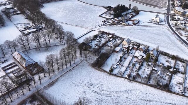 Aerial view of a snow covered rural area with scattered houses, tree lined paths, open fields, and quiet winter roads forming a calm countryside landscape.