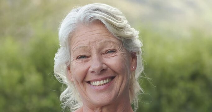 Senior female responding to lens, turning, widening smile in garden, wearing studs and light top
