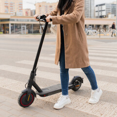 A young woman in a beige coat walks with an electric scooter through a pedestrian crossing in the city. Eco-friendly transport.