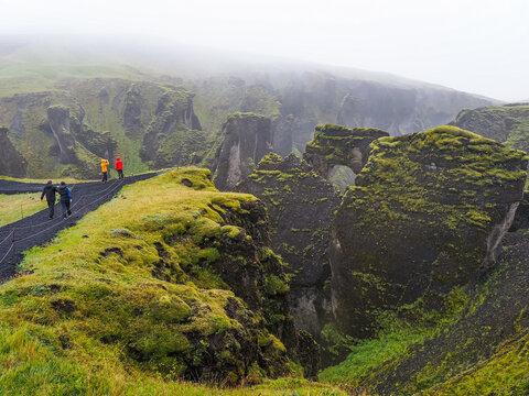 Rainy and cloudy day. People walk on winding black gravel track to the Fja&eth;r&aacute; river valley "Feather river canyon" or Fja&eth;r&aacute;rglj&uacute;fur canyon with deep moss-covered cliffs. Kirkjub&aelig;jarklaustur. Iceland.