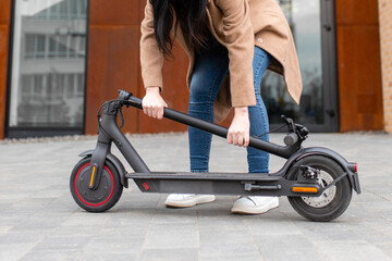 Young woman in a beige coat folds an electric scooter in the middle of a city street. A person uses an environmentally friendly form of transport in the form of an electric scooter.