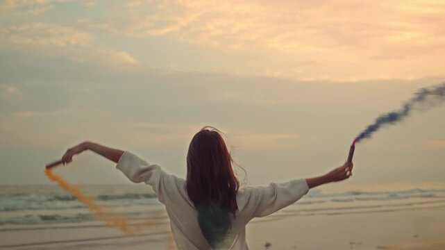 Happy Asian woman holding and playing with colorful smoke bombs on the beach during sunset. Young female enjoying summer vacation with blue and orange smoke flares. Creative celebration and freedom