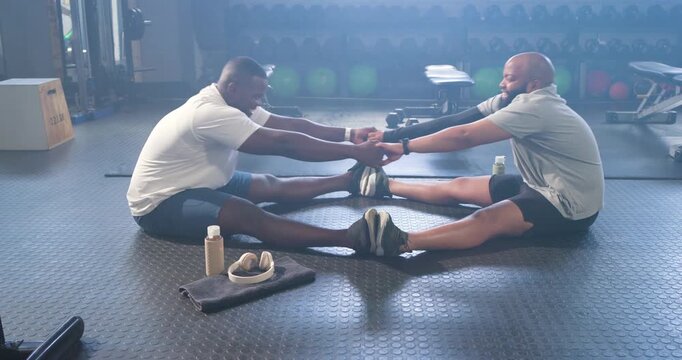 African American men in shorts holding hands on gym floor, assisting hamstring stretch with towel