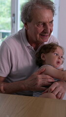 Obraz premium Grandfather holding toddler on lap at dining table, both sitting closely together in tender moment of intergenerational bonding inside bright home