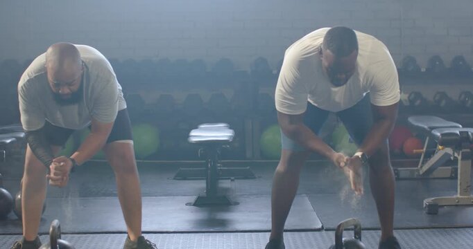 Two men clapping, creating chalk dust, hinging and reaching, gripping kettlebells at gym for swing