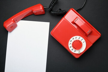 Red rotary telephone of the USSR with a handset next to a white sheet of paper on a black background.