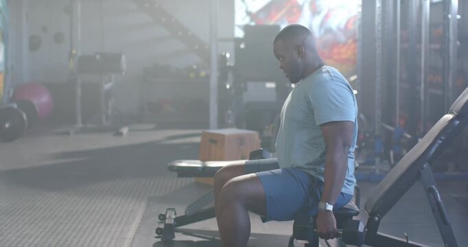 African American man glancing down, curling dumbbells on adjustable bench at gym for arm strength