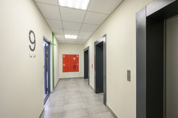 A clean, modern hallway with light beige walls, gray tiled floor, recessed ceiling lights, elevator doors, fire safety cabinet, numbered entrance (9), and green accent stripe