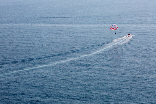 Aerial view of parasailing activity over open sea. Speedboat pulling a parasail with people flying above the water, leaving a long wake trail on the ocean surface. Travel, summer vacation, adventure a