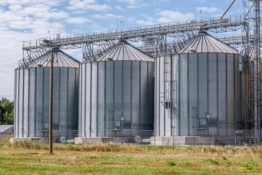 Modern agro-processing plant with silver metal silos for grain storage. Industrial granary elevator complex for drying and cleaning agricultural products against a blue cloudy sky
