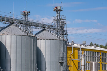Modern agro-processing plant with silver metal silos for grain storage. Industrial granary elevator complex for drying and cleaning agricultural products against a blue cloudy sky.