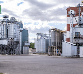 Industrial grain elevator and agro-processing plant with metal silos and conveyor system. Agricultural facility for drying, cleaning, and storage of crops under a cloudy sky.