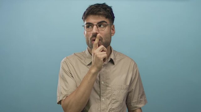 Hispanic man wearing round glasses and beige shirt standing finger to lips for silence in light blue studio; secrecy.