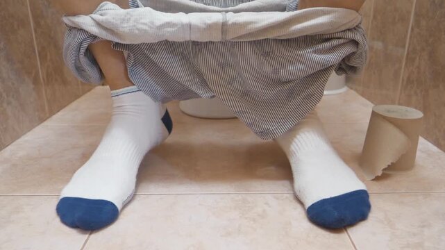 Man sits on a toilet sink in a restroom and impatiently twitches his legs. Feet of a man sitting on a toilet in white socks and with his underwear down. Man can not poop and suffer from constipation

