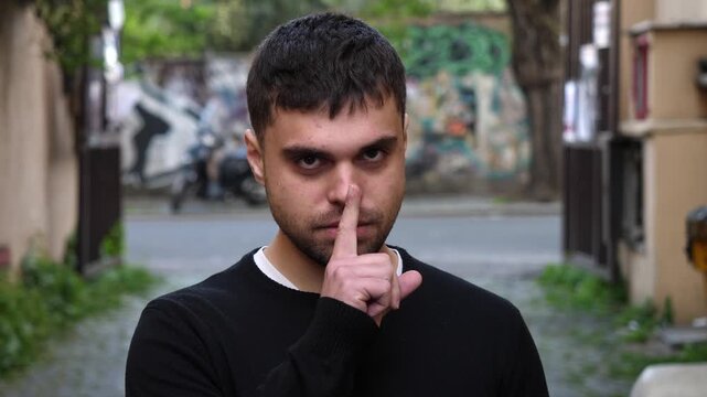 A young man asks for silence by placing his finger on his lips. Clear communication gesture expressing secrecy, calm and request for quiet attention.