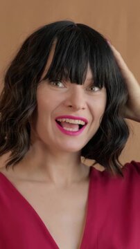 Studio Shot Of Woman Adjusting Hair And Makeup. Closeup Of Woman In Pink Dress Touching Hair And Smiling. Female Model With Red Dress And Pink Lipstick Posing With Poise And Confidence, Bangs Bob Hair