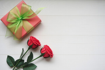 Red roses and gift box in elegant wrapping with green ribbon on white wooden background for romantic occasion