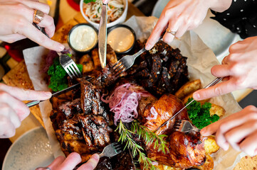 Top view of friends sharing a large grilled meat platter with ribs, roasted chicken, potatoes, and...