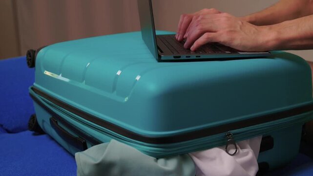 Laptop on turquoise suitcase, hands typing on keyboard on closed case improvised workspace in hotel room with clothes peeking from zip, soft evening light, focused productive mood