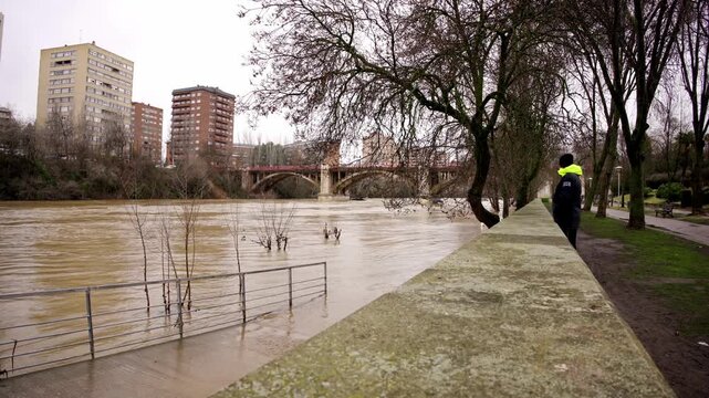 Man in a park is photographing the muddy, overflowing pisuerga river flooding the riverbanks and partially submerging trees and railings, with the city of valladolid in the background