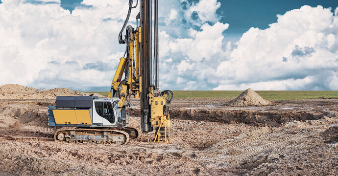 A drilling rig works on a pile foundation at a construction site with dirt and a clear sky filled with clouds in the background