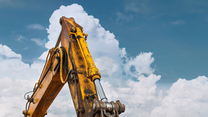 A large yellow excavator stands tall at a construction site. The machine reaches towards a clear blue sky filled with white clouds on a sunny day © Anoo