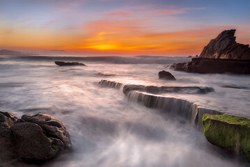 Fototapeta premium Vibrant sunset at Azkorri beach in Getxo with long exposure silky waves over mossy rocks in Spain