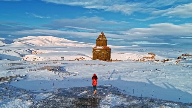 Aerial view of a tourist photographing the Abughamrents Church at Ani Ruins in Kars, Turkey. Snow covered medieval Armenian chapel stands in a vast winter landscape.