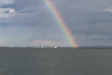 Vibrant rainbow arcs over a calm sea with a distant cityscape under cloudy skies