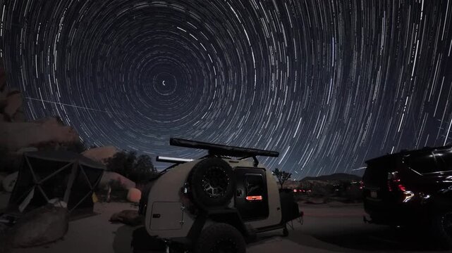 Teardrop camper sits at a campsite while star trails move across the night sky above Joshua Tree National Park in California.