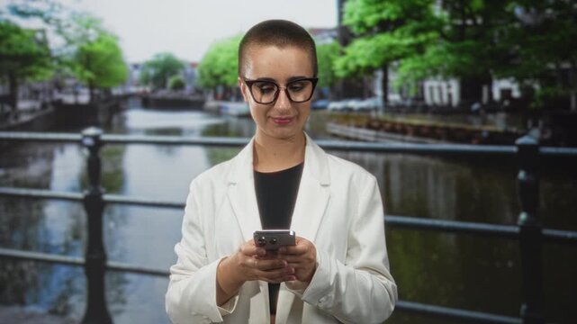 Woman with buzzcut and glasses holds smartphone with both hands while scrolling on a canal side street bridge; contemplative.