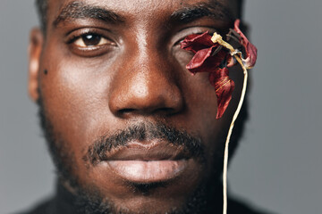 African man portrait with dried red flower near eye in creative conceptual visual style. Closeup face with dark skin and artistic expression on gray background for modern art.