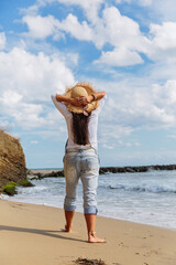 Mature woman stands with arms behind head holding straw hat and sunglasses looking out at sea