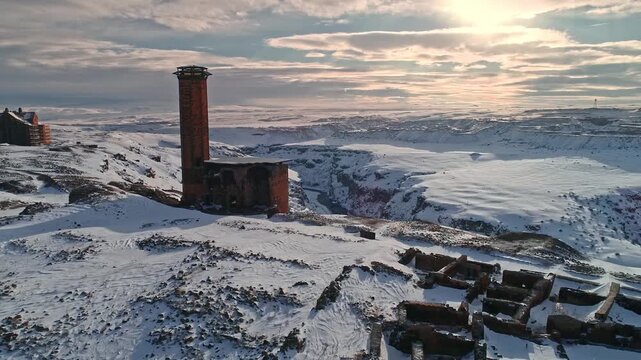 Aerial view of the Menucehr Mosque minaret at Ani Ruins in Kars, Turkey during sunset. Snow covered medieval architecture stands against a glowing winter sky.
